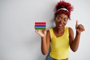 mujer africana con cabello afro, use camiseta amarilla y anteojos, sostenga la bandera de gambia aislada en fondo blanco, muestre el pulgar hacia arriba. foto