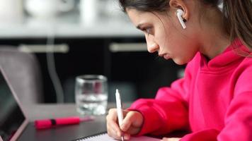 mujer joven con una sudadera rosa brillante sentada en una mesa con auriculares de tableta mientras escribe en un cuaderno con un bolígrafo video