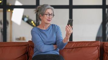mulher com cabelos grisalhos e óculos se senta em um sofá usando um telefone inteligente para uma chamada de vídeo video