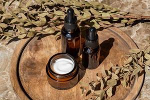 suero facial de colágeno antienvejecimiento en botella de vidrio oscuro y crema facial en bandeja redonda de madera con ramo de eucalipto seco. concepto de belleza cosmética orgánica natural. foto