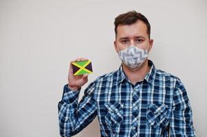 Man in checkered shirt show Jamaica flag card in hand, wear protect mask isolated on white background. American countries Coronavirus concept. photo