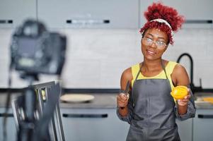 African american woman filming her blog broadcast about healthy food at home kitchen. She cuts a tomato with knife. photo