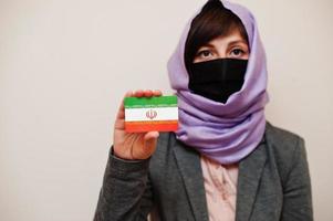 retrato de una joven musulmana que usa ropa formal, protege la máscara facial y el pañuelo de la cabeza del hiyab, sostiene la tarjeta de la bandera de Irán contra un fondo aislado. concepto de país coronavirus. foto