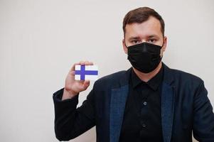 el hombre europeo usa negro formal y protege la máscara facial, sostiene la tarjeta de la bandera de finlandia aislada en el fondo blanco. concepto de país covid coronavirus de europa. foto