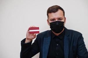 el hombre europeo usa negro formal y protege la máscara facial, sostiene la tarjeta de la bandera de letonia aislada en el fondo blanco. concepto de país covid coronavirus de europa. foto