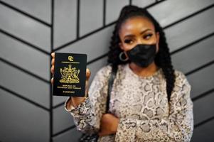 African american woman wearing black face mask show Trinidad and Tobago passport in hand. Coronavirus in America country, border closure and quarantine, virus outbreak concept. photo