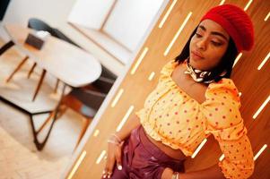 Elegant afro american woman in red french beret, big gold neck chain polka dot blouse and leather pants pose indoor. photo