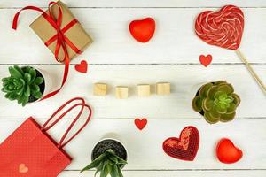 Creative Valentine Day romantic composition with red hearts, satin ribbon, lollipop, gift box and paper bag on white background. Mockup with empty wooden cubes for text. photo