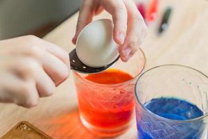 Transparent glass cups with colored water. The process of coloring eggs. A child hands put in a white egg with a spoon. photo