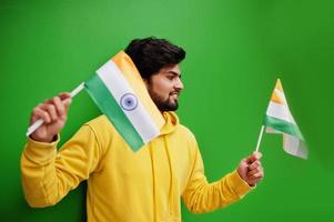 Urban young hipster indian man in a fashionable yellow sweatshirt hold flags of India in hands. Cool south asian guy wear hoodie isolated on green background. photo