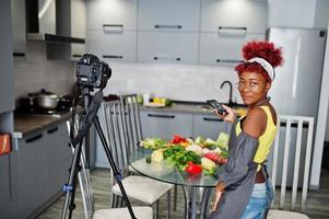 mujer afroamericana filmando la transmisión de su blog sobre comida saludable en la cocina de casa y haciendo fotos por teléfono.