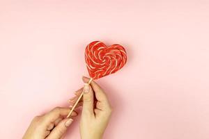 Female hand holding Heart shape lollipop on pink background. Romance flat lay with copy space photo