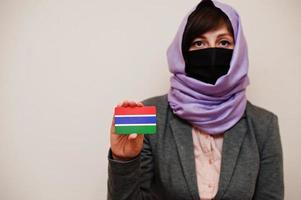 Portrait of young muslim woman wearing formal wear, protect face mask and hijab head scarf, hold The Gambia flag card against isolated background. Coronavirus country concept. photo