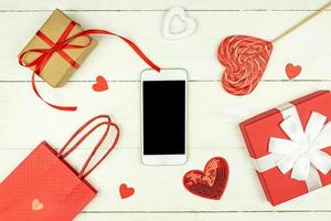 Creative Valentine Day romantic composition with red hearts, smartphone, lollipop, gift box and paper bag on pink background. Mockup with copy space for blogs and social media photo