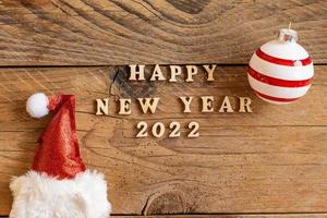 feliz año nuevo 2022 cita de letras de madera con sombrero rojo brillante de santa y adorno de rayas en la mesa de madera. tarjeta de felicitación festiva para una celebración de año nuevo foto