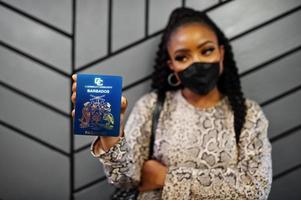 mujer afroamericana con mascarilla negra muestra el pasaporte de barbados en la mano. coronavirus en el país de América, cierre de fronteras y cuarentena, concepto de brote de virus. foto