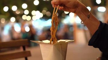 Close up of feminine hand using chopsticks to pick up noodles from a chinese take out box at an outdoor table video