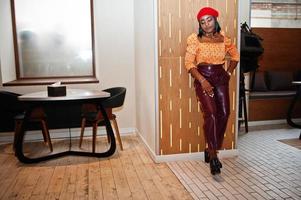 Elegant afro american woman in red french beret, big gold neck chain polka dot blouse and leather pants pose indoor. photo