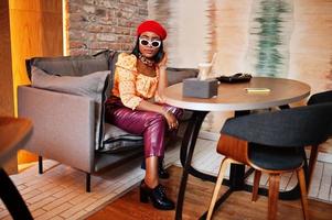 Elegant afro american woman in red french beret, big gold neck chain polka dot blouse and leather pants sitting at restaurant and drink latte. photo
