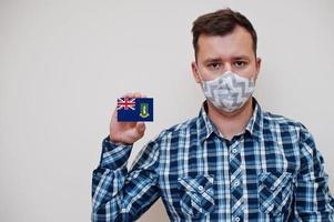Man in checkered shirt show British Virgin Islands flag card in hand, wear protect mask isolated on white background. American countries Coronavirus concept. photo