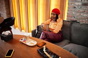 Elegant afro american woman in red french beret, big gold neck chain polka dot blouse and leather pants sitting at restaurant and drink latte. photo