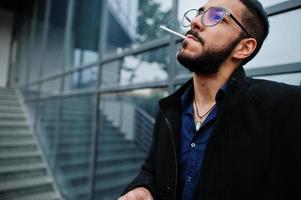 Middle eastern entrepreneur wear black coat and blue shirt, eyeglasses against office building smoking cigarette. photo