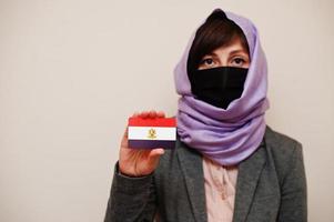 Portrait of young muslim woman wearing formal wear, protect face mask and hijab head scarf, hold Egypt flag card against isolated background. Coronavirus country concept. photo