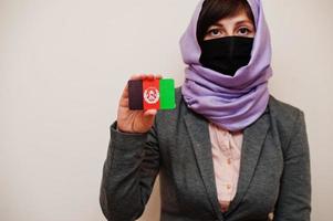 Portrait of young muslim woman wearing formal wear, protect face mask and hijab head scarf, hold Afghanistan flag card against isolated background. Coronavirus country concept. photo