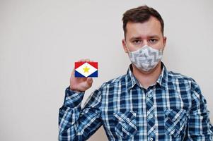 el hombre con camisa a cuadros muestra la tarjeta de la bandera de saba en la mano, usa una máscara protectora aislada en el fondo blanco. concepto de coronavirus de los países americanos. foto