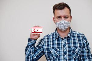 Man in checkered shirt show Northern Cyprus flag card in hand, wear protect mask isolated on white background. Europe countries Coronavirus concept. photo