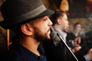 Group of handsome retro well-dressed man gangsters spend time at club, smoking hookah. Multiethnic male bachelor mafia party in restaurant. photo