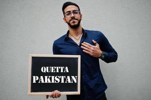 el hombre árabe usa camisa azul y anteojos sostiene el tablero con la inscripción quetta pakistan. ciudades más grandes en el concepto del mundo islámico. foto