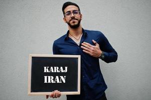 el hombre árabe usa camisa azul y anteojos sostiene una tabla con la inscripción karaj iran. ciudades más grandes en el concepto del mundo islámico. foto
