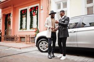 Two fashion black men stand near business car and look at cell phone. Fashionable portrait of african american male models. Wear suit, coat and hat. photo