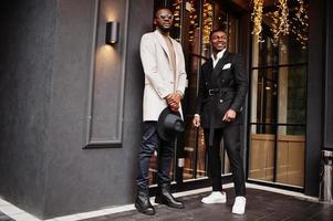 Two fashion black men pose against house with garlands. Fashionable portrait of african american male models. Wear suit, coat and hat. photo