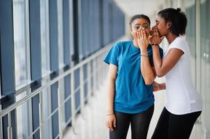 Two african woman friends in t-shirts whispers gossip indoor together. photo