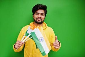 Urban young hipster indian man in a fashionable yellow sweatshirt hold flags of India in hands. Cool south asian guy wear hoodie isolated on green background. photo