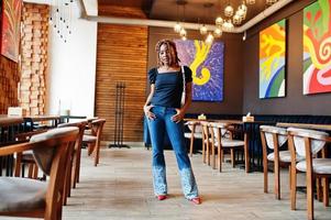 Lovely african american woman with dreadlocks in blue stylish jeans at cafe. Beautiful cool fashionable black young girl indoor. photo