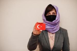 retrato de una joven musulmana que usa ropa formal, protege la máscara facial y el pañuelo de la cabeza del hiyab, sostiene la tarjeta de la bandera de turquía contra un fondo aislado. concepto de país coronavirus. foto