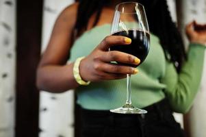 Cierra las manos de una atractiva joven afroamericana vestida con suéter verde y jeans negros posan en el restaurante con una copa de vino tinto.