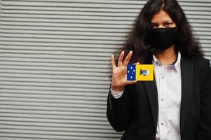 Asian woman at formal wear and black protect face mask hold Australian Capital Territory flag at hand against gray background. Coronavirus Australia state concept. photo