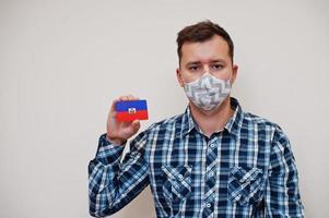 el hombre con camisa a cuadros muestra la tarjeta de la bandera de haití en la mano, usa una máscara protectora aislada en el fondo blanco. concepto de coronavirus de los países americanos. foto