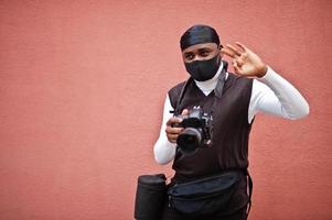 joven camarógrafo afroamericano profesional con cámara profesional con equipo profesional. camarógrafo afro con duraq negro y máscara de protección facial, haciendo videos. foto