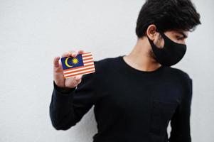 Asian man wear all black with face mask hold Malaysia flag in hand isolated on white background. Coronavirus country concept. photo