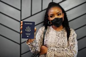 African american woman wearing black face mask show USA passport in hand. Coronavirus in America country, border closure and quarantine, virus outbreak concept. photo