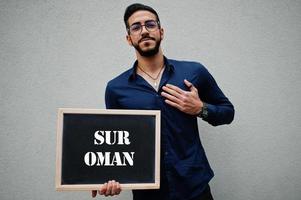 Arab man wear blue shirt and eyeglasses hold board with Sur Oman inscription. Largest cities in islamic world concept. photo