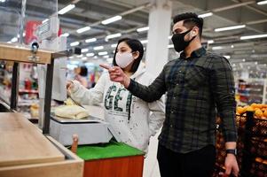 Asian couple wear in protective face mask shopping together in supermarket during pandemic. Weigh the goods. photo