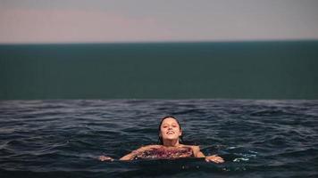 Beautiful woman swims in a pool of dark blue water and raises up at edge to look out video