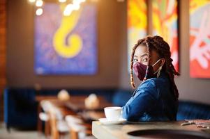 Stylish african american woman with dreadlocks afro hair, wear jeans jacket and face protect  mask at restaurant. New normal life after coronavirus epidemic.