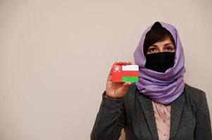 Portrait of young muslim woman wearing formal wear, protect face mask and hijab head scarf, hold Oman flag card against isolated background. Coronavirus country concept. photo